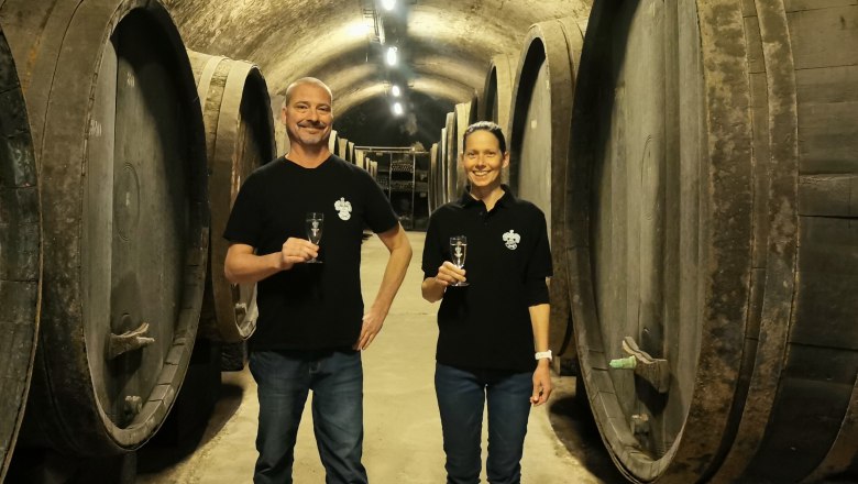 Die Eigentümer der Sektkellerei Esther Inführ und Thomas Schlatte im historischen Fasskeller, © Sektkellerei Inführ Zwei Personen stehen in einem Weinkeller mit großen Holzfässern und halten Sektgläser.