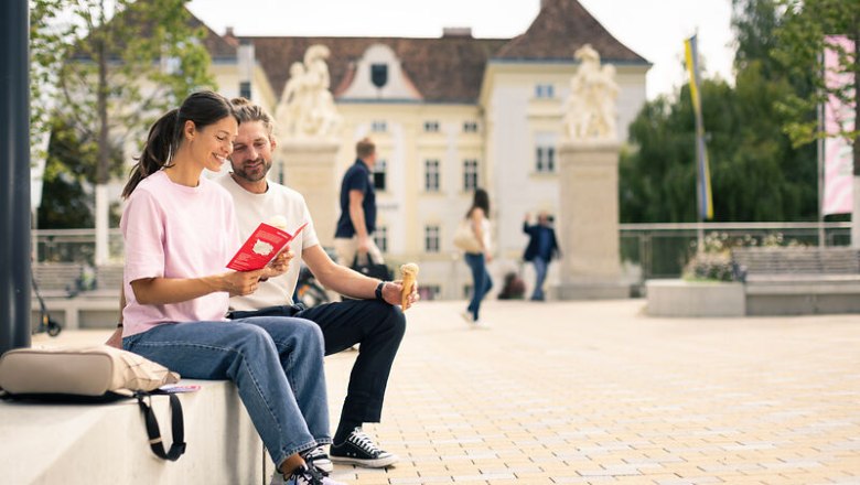 Schloss Bad Vöslau, © Wienerwald Tourismus/Martin Fülöp Ein Paar sitzt auf einer Bank im Freien und liest gemeinsam ein Buch. Im Hintergrund ist ein historisches Gebäude zu sehen.