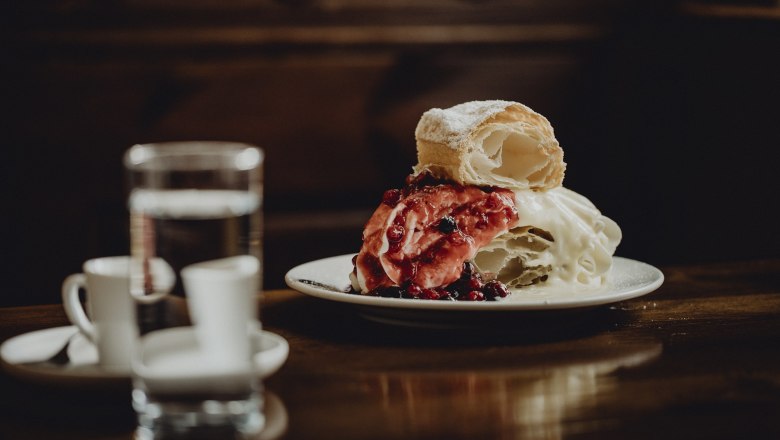 Klostergasthof Heiligenkreuz, © Niederösterreich Werbung/Sophie Menegaldo Ein Teller mit Strudel und Beeren, daneben ein Glas Wasser und zwei Espressotassen auf einem Holztisch.