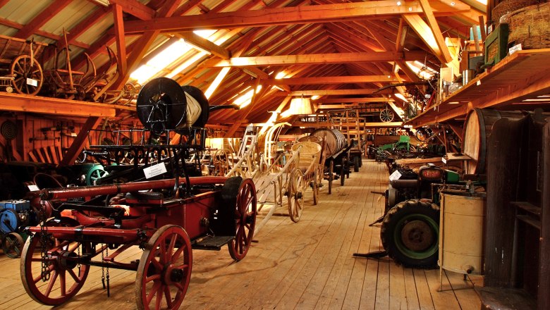 Innenansicht eines Museums mit alten landwirtschaftlichen Ger&auml;ten und Wagen auf Holzboden.