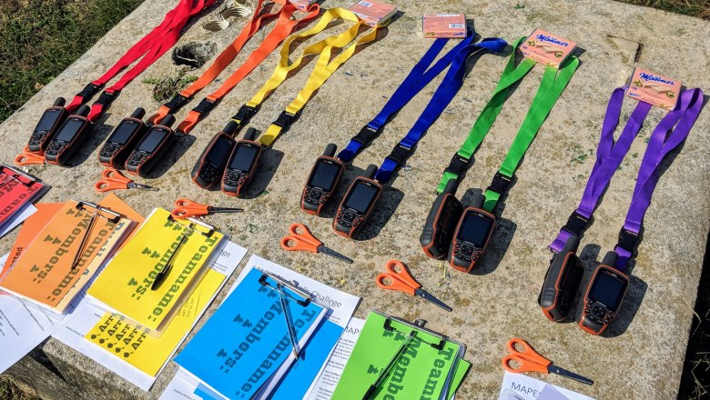 Bunt vorbereitete Teambuilding-Station mit GPS-Ger&auml;ten, farbigen Team-B&auml;ndern und Aufgabenmappen f&uuml;r eine spannende Outdoor-Challenge in der Natur.