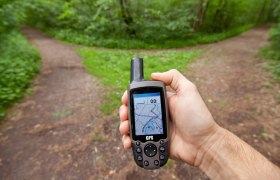 Eine Hand h&auml;lt ein GPS-Ger&auml;t vor einem Weg im Wald, der sich in zwei Richtungen teilt.
