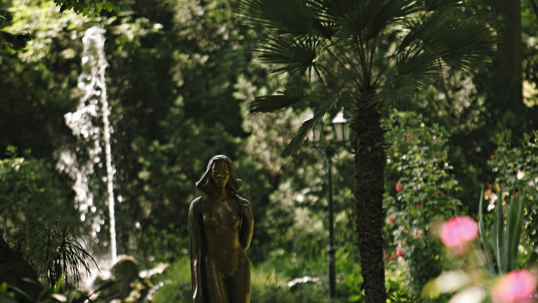 Eine Statue in einem gr&uuml;nen Garten mit einem Springbrunnen im Hintergrund.