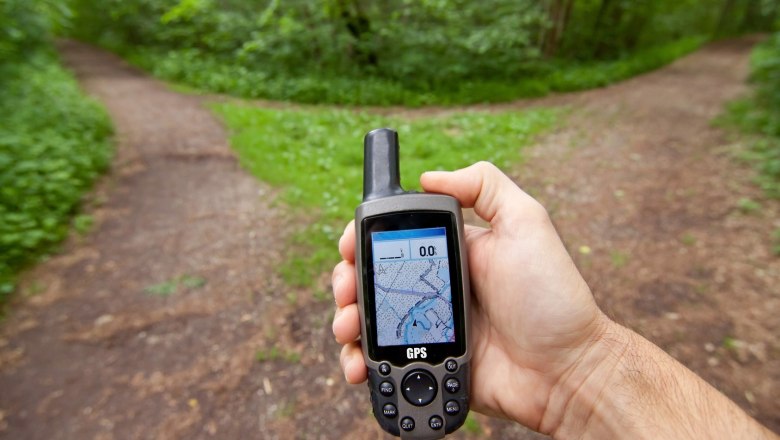 Eine Hand h&auml;lt ein GPS-Ger&auml;t vor einem Weg im Wald, der sich in zwei Richtungen teilt.